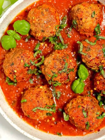 meatballs in a bowl of red sauce with fresh herbs on top and red pepper flakes