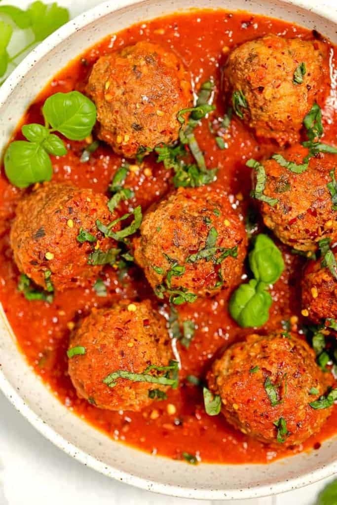 meatballs in a bowl of red sauce with fresh herbs on top and red pepper flakes