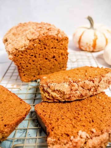 pumpkin bread loaf and pumpkin bread slices laying on top of eachother with a crumble topping and a pumpkin in the background