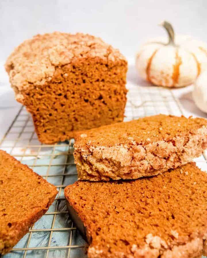 pumpkin bread loaf and pumpkin bread slices laying on top of eachother with a crumble topping and a pumpkin in the background
