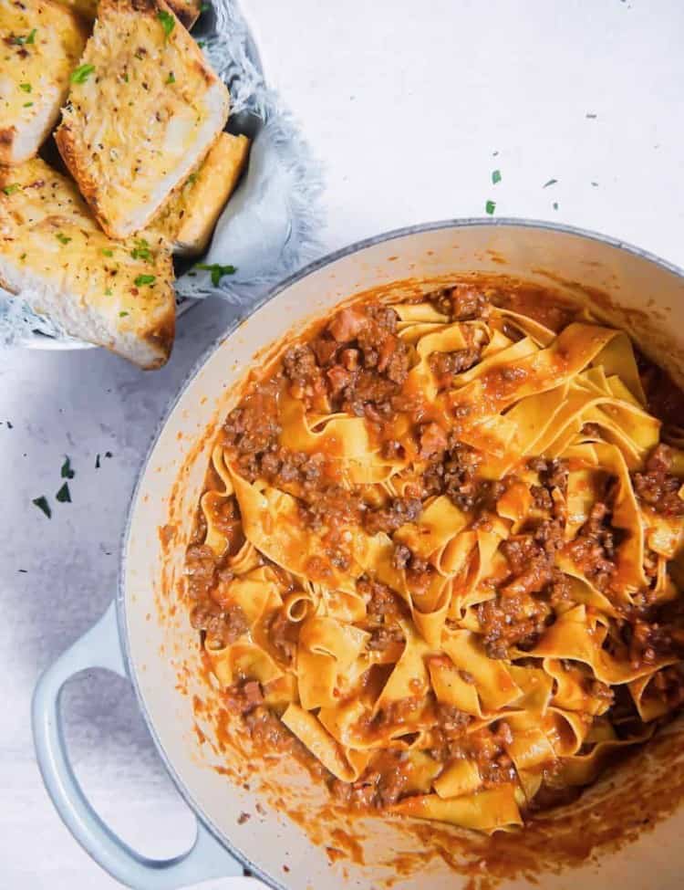 an overhead shot of bolognese sauce mixed with pappardelle pasta and garlic bread in a bowl to the side