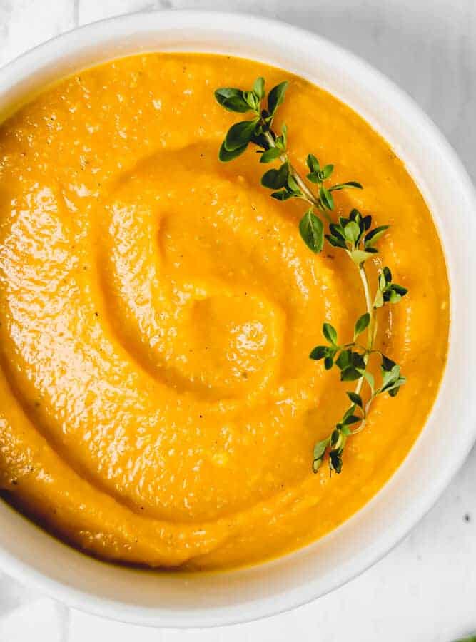 close up of butternut squash soup in a white bowl with a fresh thyme sprig