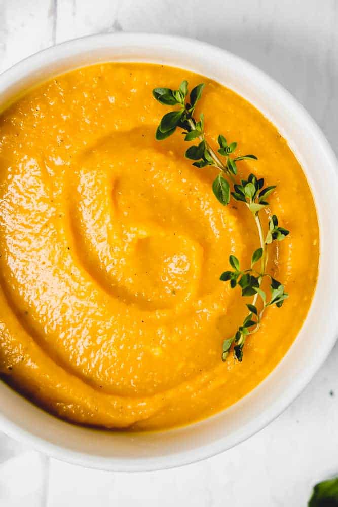 close up of butternut squash soup in a white bowl with a fresh thyme sprig