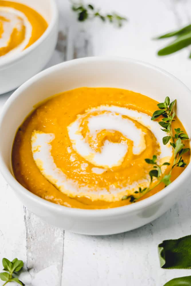 white bowl with butternut squash soup, a swirl of coconut cream, topped with fresh thyme 