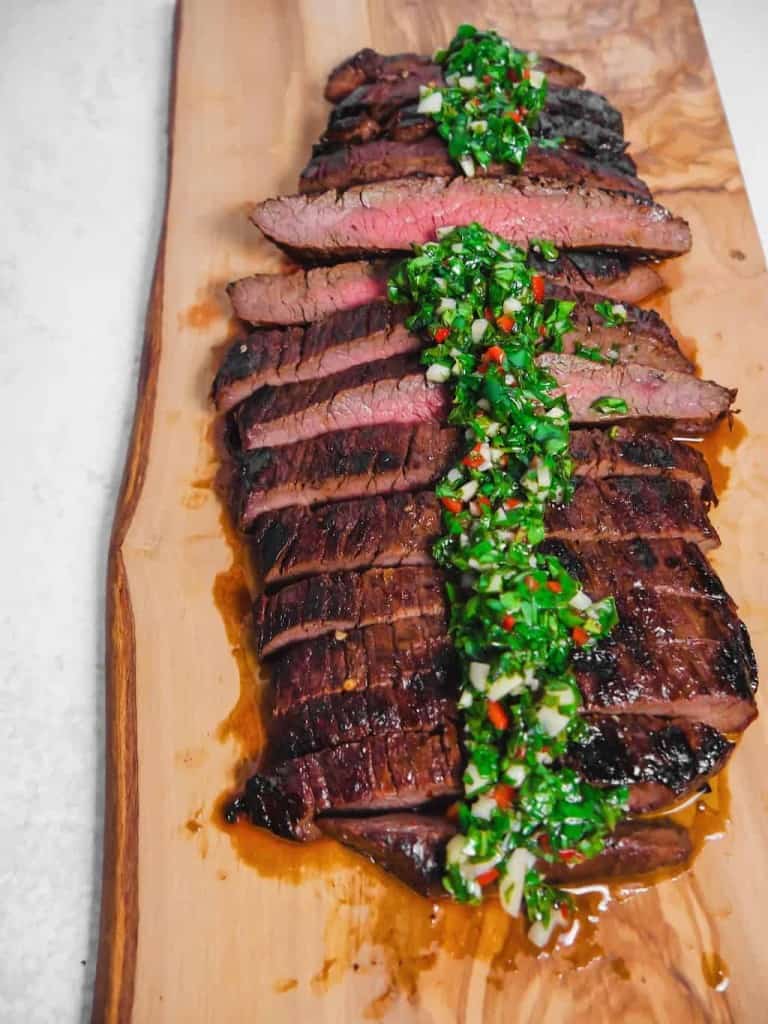 sliced grilled flank steak with chimichurri over the top on a cutting board
