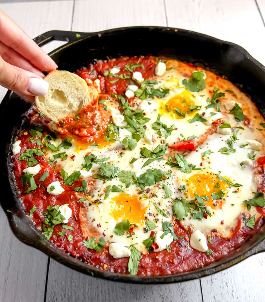 dipping a slice of bread into the shakshuka in a cast iron skillet