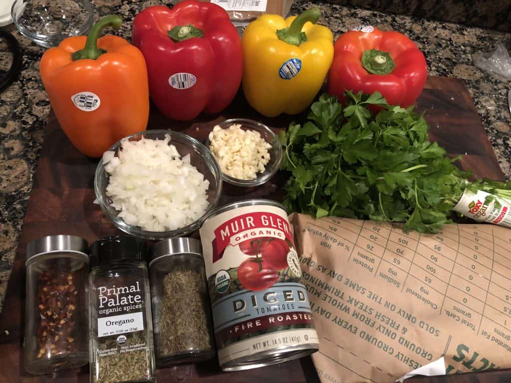stuffed pepper ingredients uncooked on cutting board. Bell peppers lined up in back, spices in jars, ad herbs