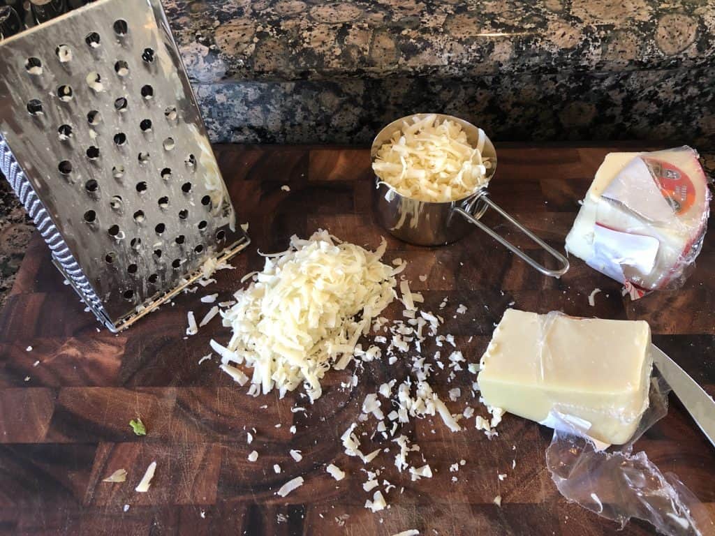 shot of shredded gruyere  and fontina cheeses on a cutting board, a cheese grater, and measuring cup on a wooden cutting board