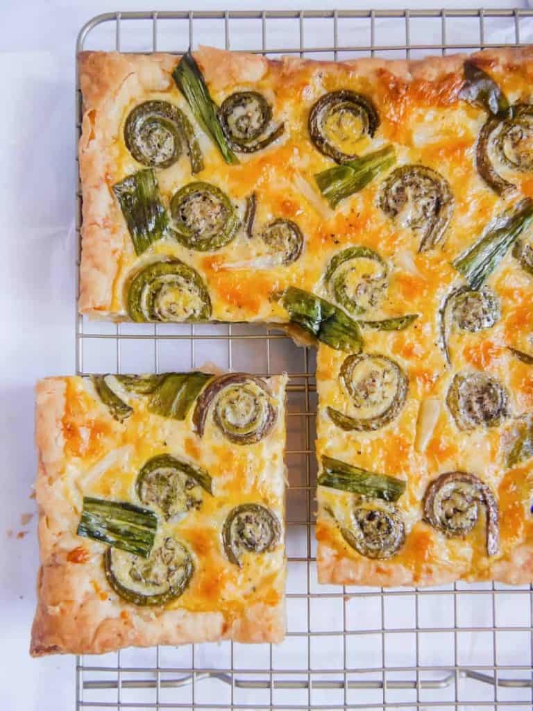 overhead shot of a square puff pastry cheese tart with roasted fiddleheads, ramps with a piece cut out on a metal wire rack