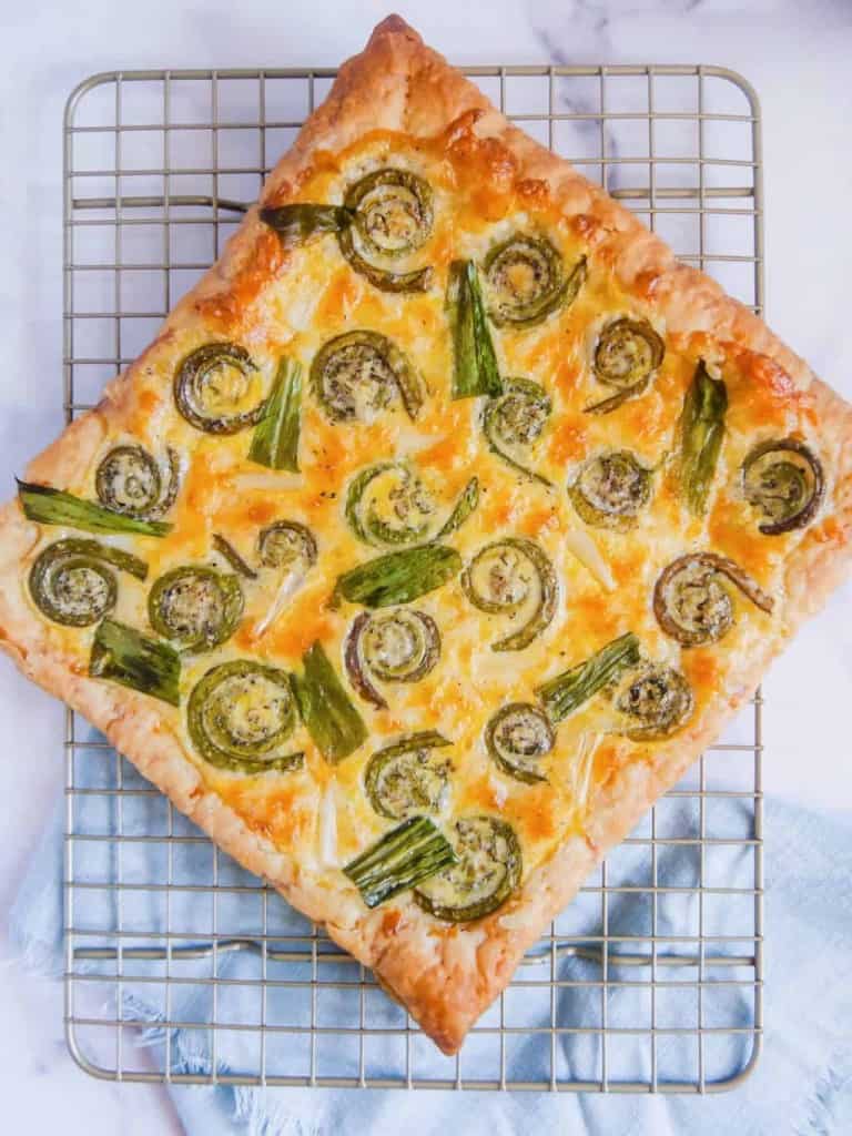 overhead shot of cooked puff pastry cheese tart with roasted fiddlehead ferns and ramps on a metal rack