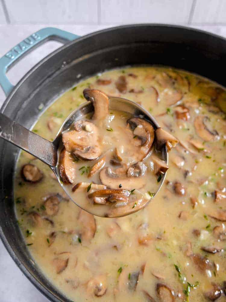 soup pot filled with mushroom soup and herbs with a ladle lifting soup out of pot