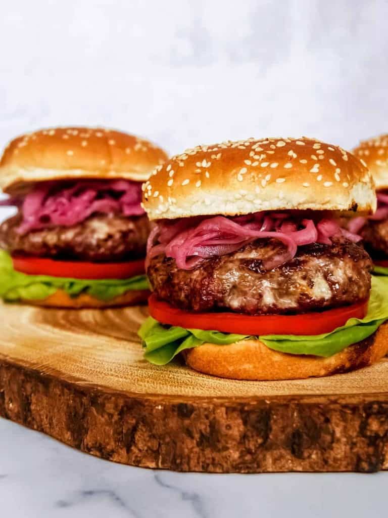 close up of blue cheese burger with pickled red onion, lettuce, tomato, sesame seed buns, stacked on wood block
