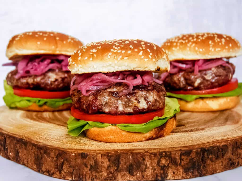 front shot of three blue cheese burgers next to each other garnished with pickled red onions, sliced tomatoes, and lettuce on a wooden block