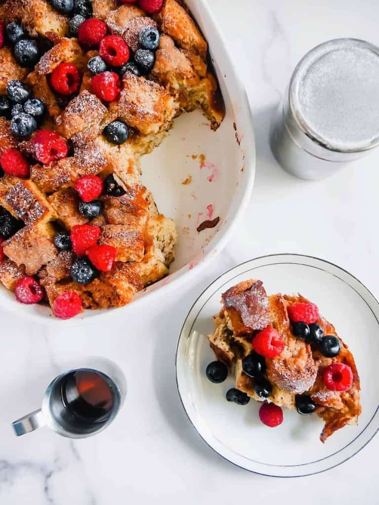 over head shot of french toast casserole with berries in a baking dish with section missing, a plate with a serving of french toast, maple syrup container, and powdered sugar cup