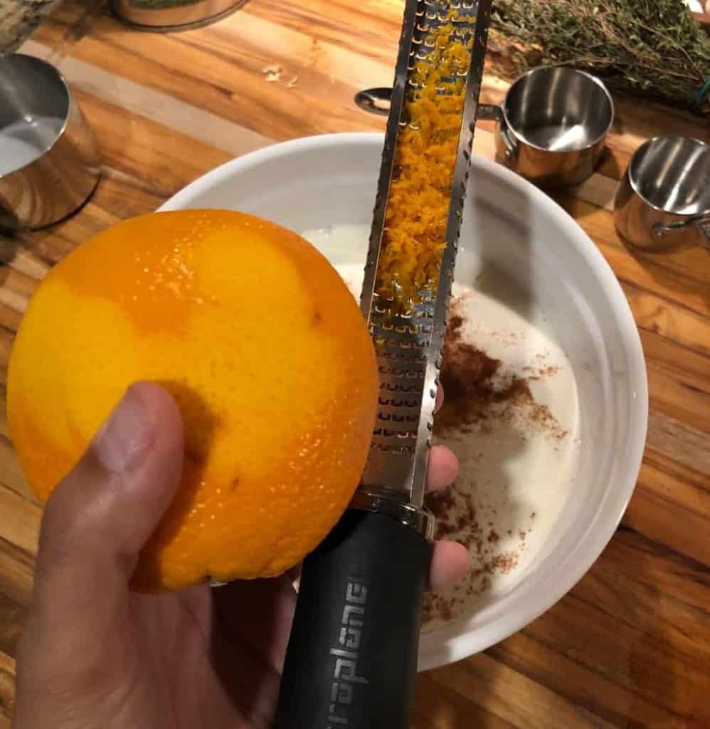 microplane with orange zest shavings and orange in hand with white bowl underneath 