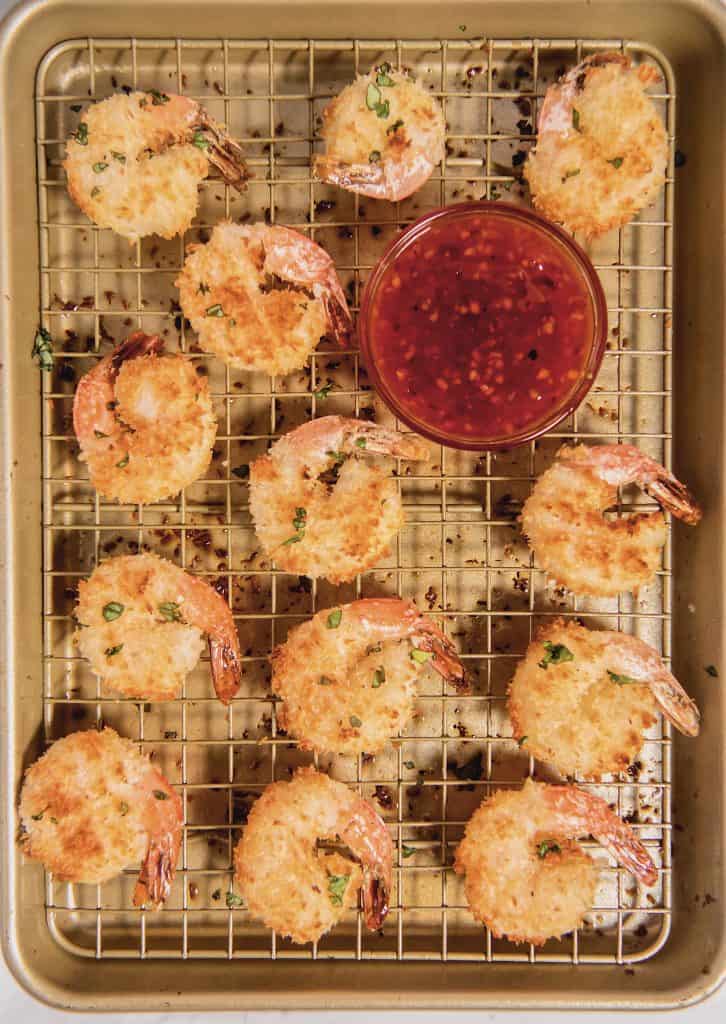 overhead shot of baked coconut shrimps on a baking rack with sweet chili sauce in a bowl in the middle