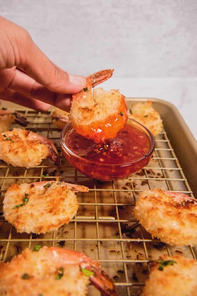 close up of a baked coconut shrimp being dipped into the sweet spicy chili sauce and other shrimps on a baking rack