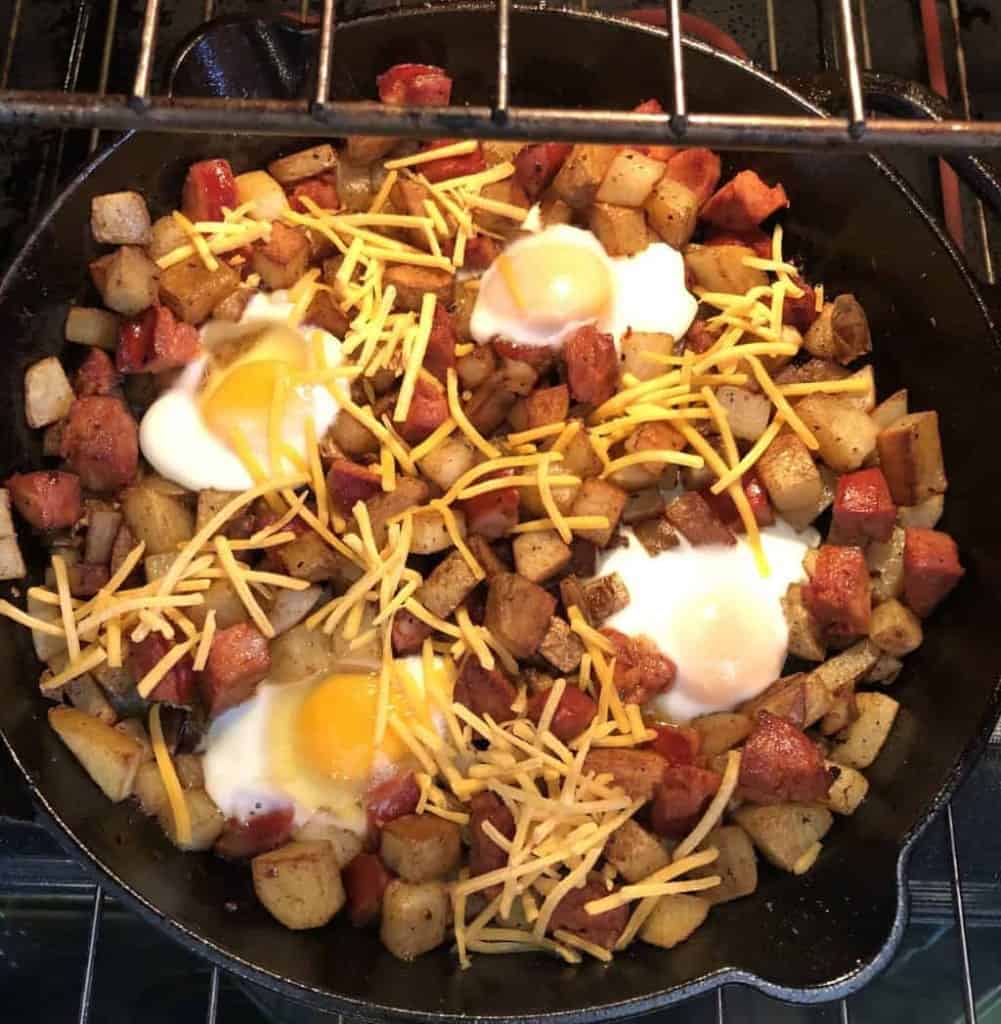 cast iron skillet in the oven with potatoes, chorizo, half cooked eggs, and shredded cheddar cheese on top