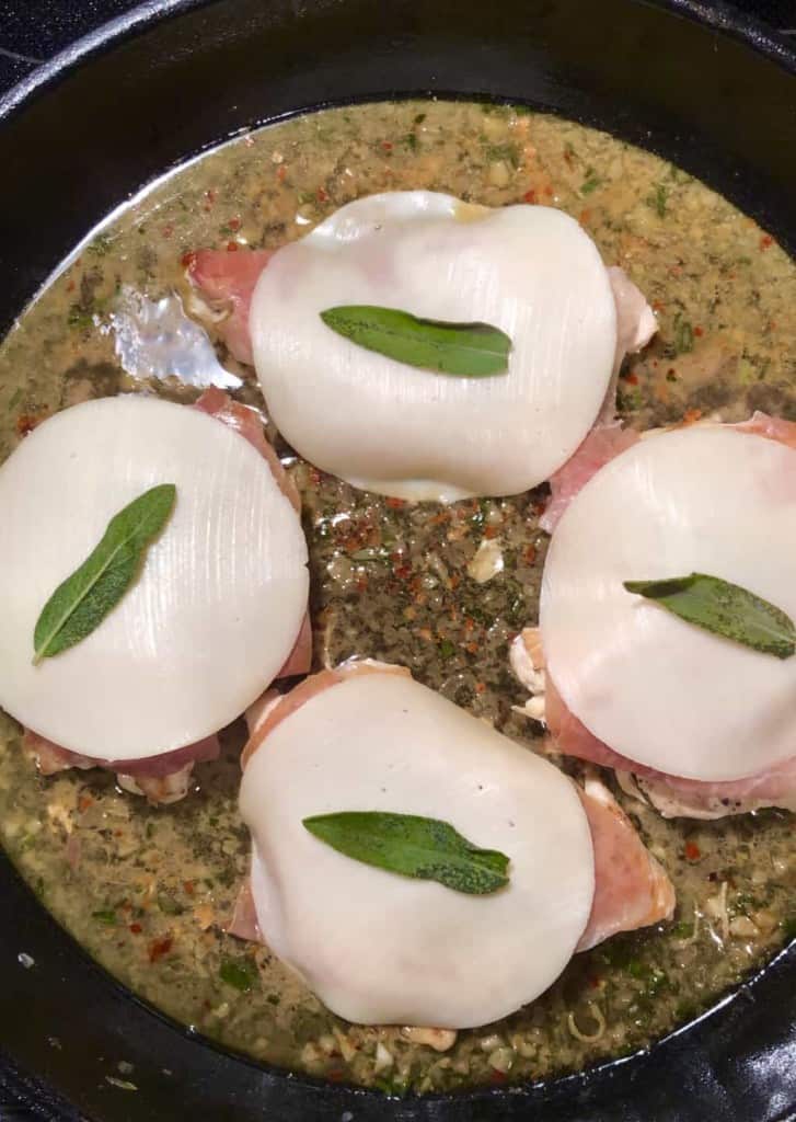 cast iron skillet with four chicken breasts topped with cheese and sage leaves