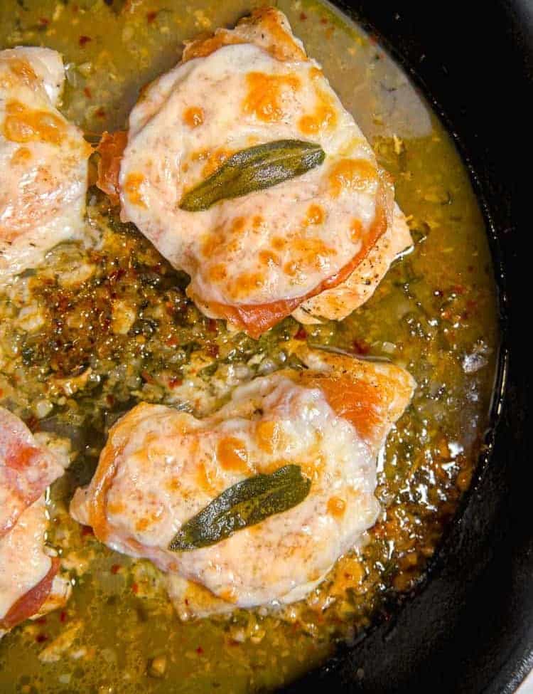 cast iron skillet with 3 pieces of chicken covered in cheese, prosciutto, and a sage leaf