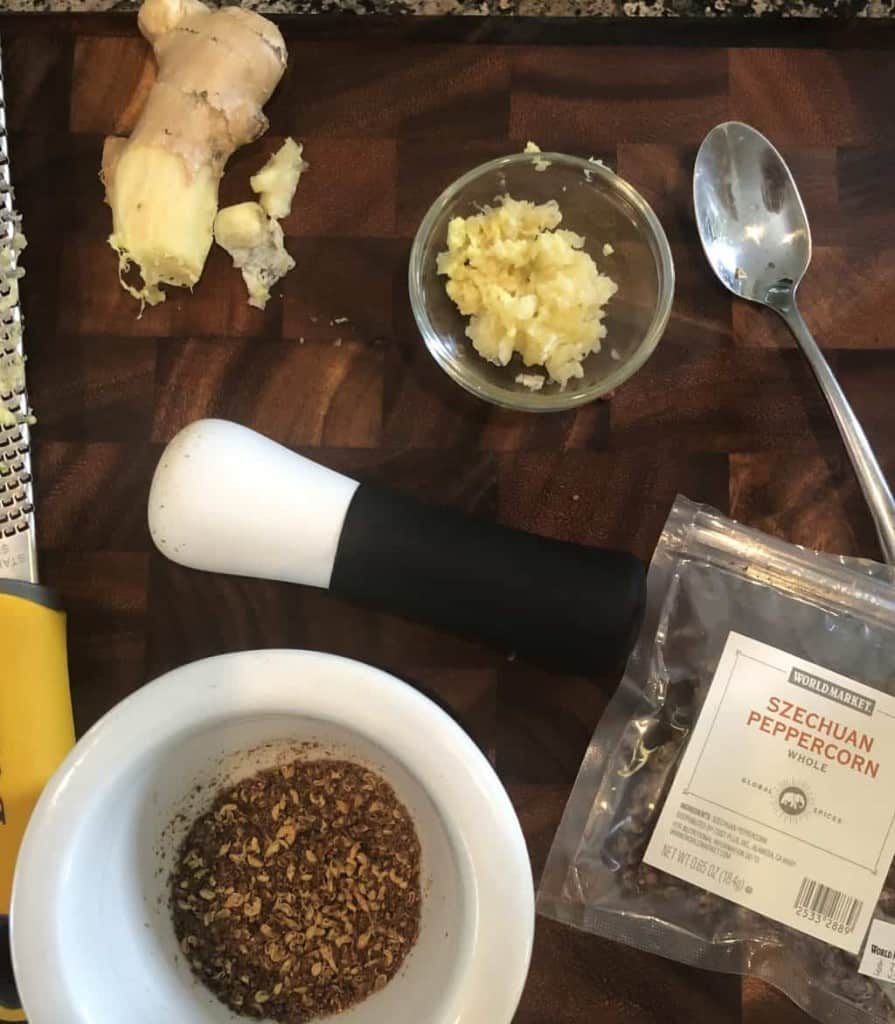 Szechuan peppercorns in a mortar and pestle, grated ginger and garlic, on a wooden cutting board