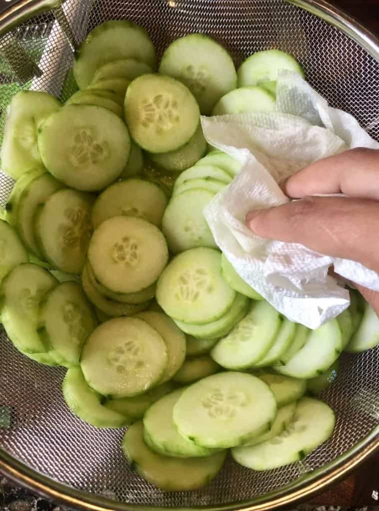 sliced cucumbers in a colander being pat dry with a paper towel 