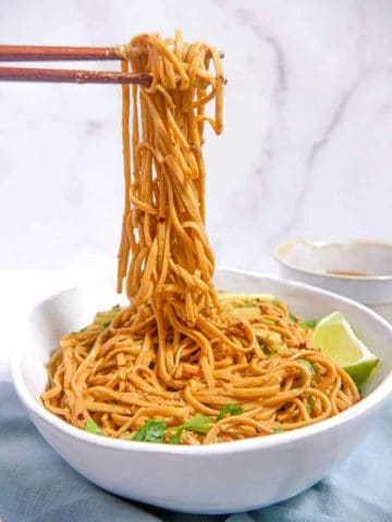 a white bowl full of sesame noodles, chopsticks lifting noodles out of the bowl into the air