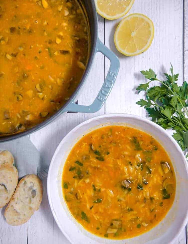 pot with vegetable orzo soup next to bowl filled with soup and bread, herbs, and lemons in background