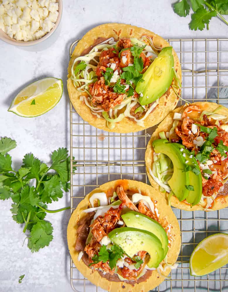 overhead photo of chicken tinga on corn tortillas, refried beans, lettuce, avocadoes on a rack with limes