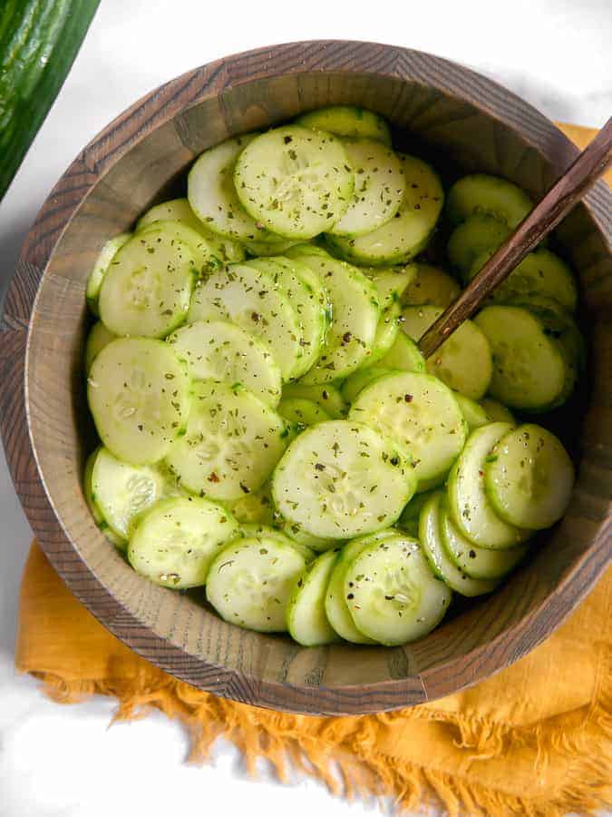 overhead shot of sliced cucumbers in a wooden bowl with a spoon in the center, laying on top of a gold towel