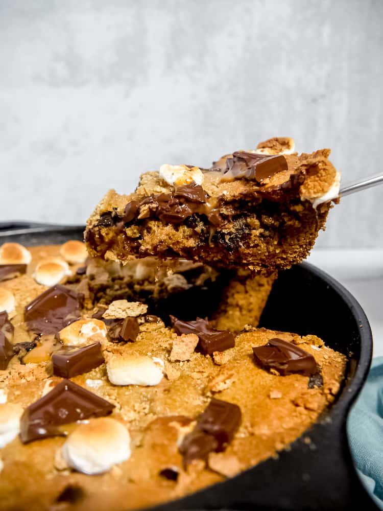 slice being pulled out of a large s'mores skillet cookie with melted chocolate, toasted marshmallows, and graham crackers on top