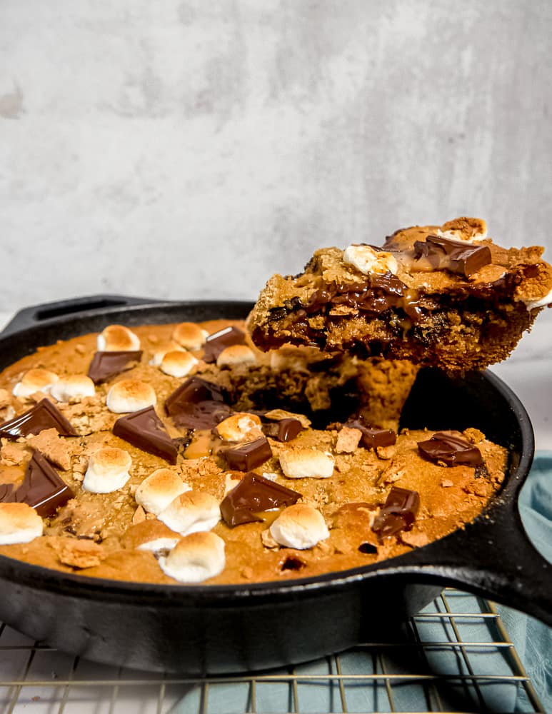 slice being pulled out of a large s'mores cookie in a cast iron skillet with melted chocolate, toasted marshmallows, and graham crackers on top