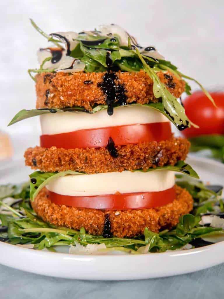 close up of a stack of crispy eggplant, tomato, mozzarella, arugula salad, piled high on a white plate with a red tomato, basil, and parmesan with balsamic glaze dripping off
