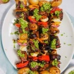 Steak kabobs with potatoes, peppers, onions, and garlic cloves, on a skewer with chimichurri sauce drizzled oer the top on a white plate