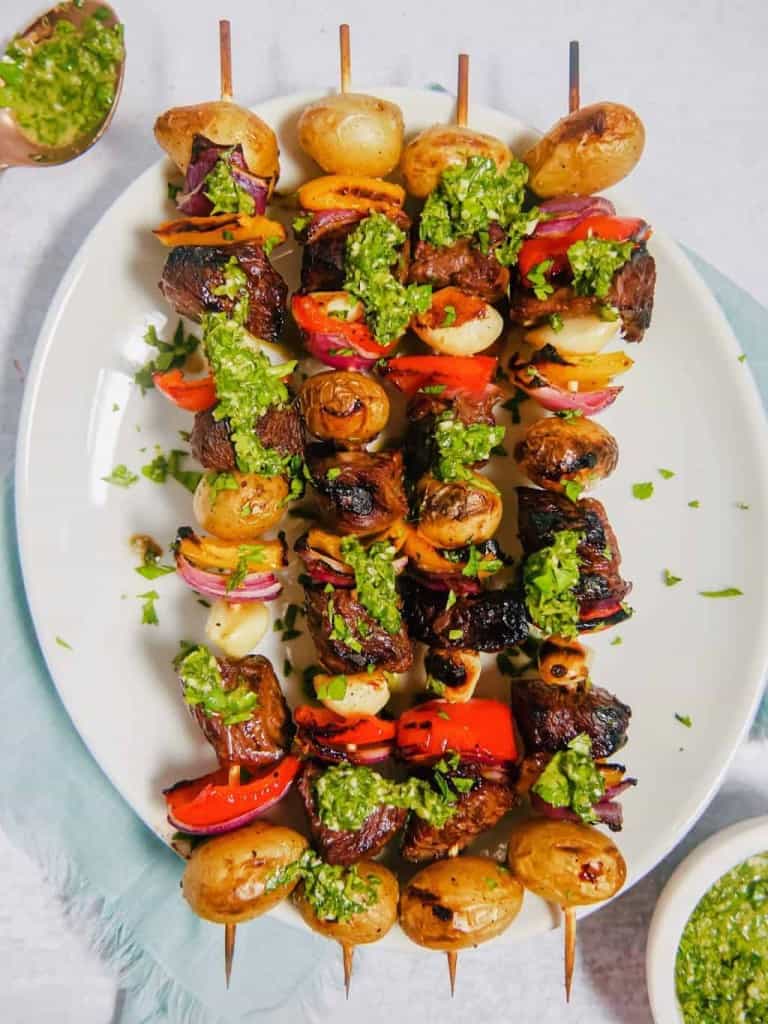 Steak kabobs with potatoes, peppers, onions, and garlic cloves, on a skewer with chimichurri sauce drizzled oer the top on a white plate