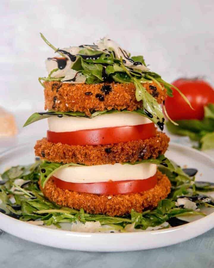 a stack of crispy eggplant, tomato, mozzarella, arugula salad, piled high on a white plate with a red tomato, basil, and parmesan cheese block in the background
