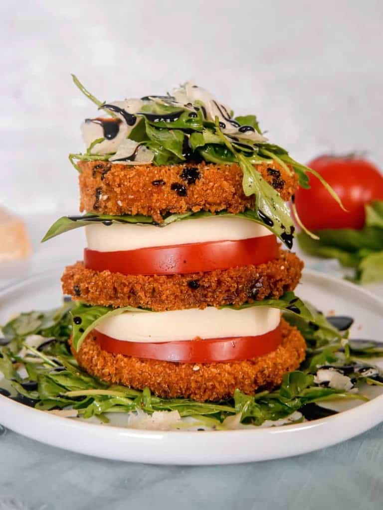 a stack of crispy eggplant, tomato, mozzarella, arugula salad, piled high on a white plate with a red tomato, basil, and parmesan cheese block in the background