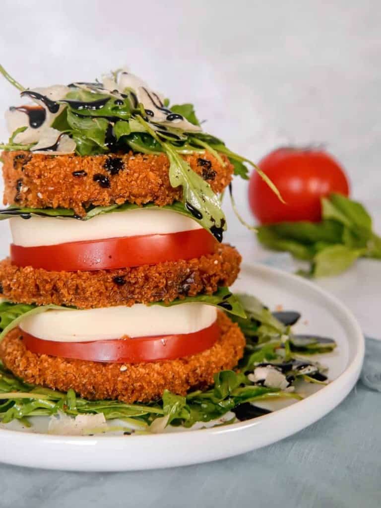 close up of a stack of crispy eggplant, tomato, mozzarella, arugula salad, piled high on a white plate with a red tomato, basil, and parmesan cheese block in the background