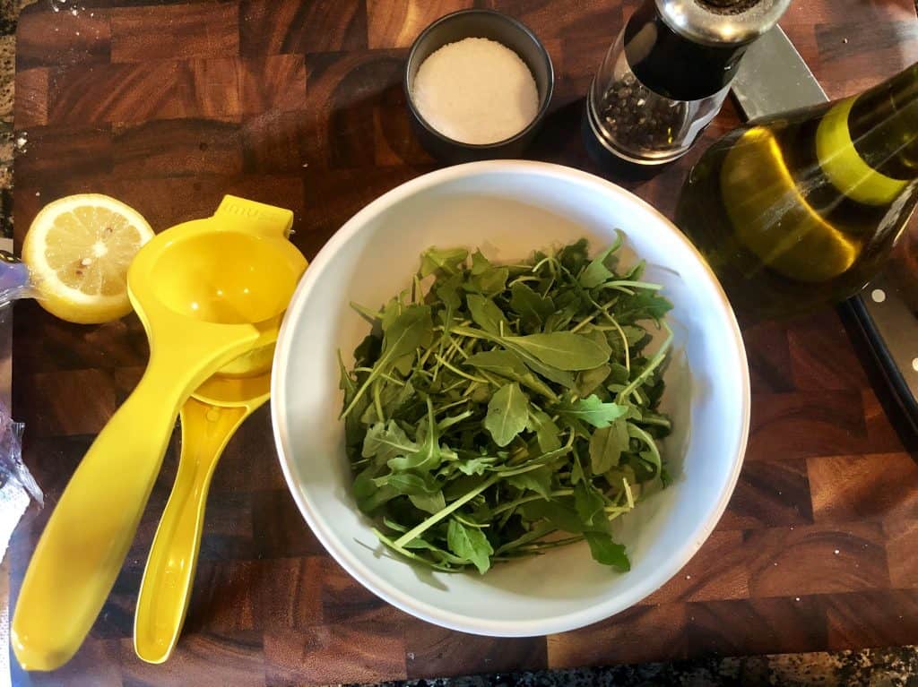 arugula in a bowl, lemon squeeze, olive oil, salt and pepper on a cutting board