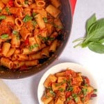 Red dutch oven filled with rigatoni, bell pepper, and eggplant, a white bowl filled with pasta and basil leaves on top