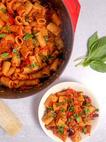 Red dutch oven filled with rigatoni, bell pepper, and eggplant, a white bowl filled with pasta and basil leaves on top