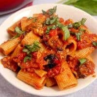 white bowl with rigatoni, red sauce, roasted eggplant an roasted bell peppers topped with basil chopped