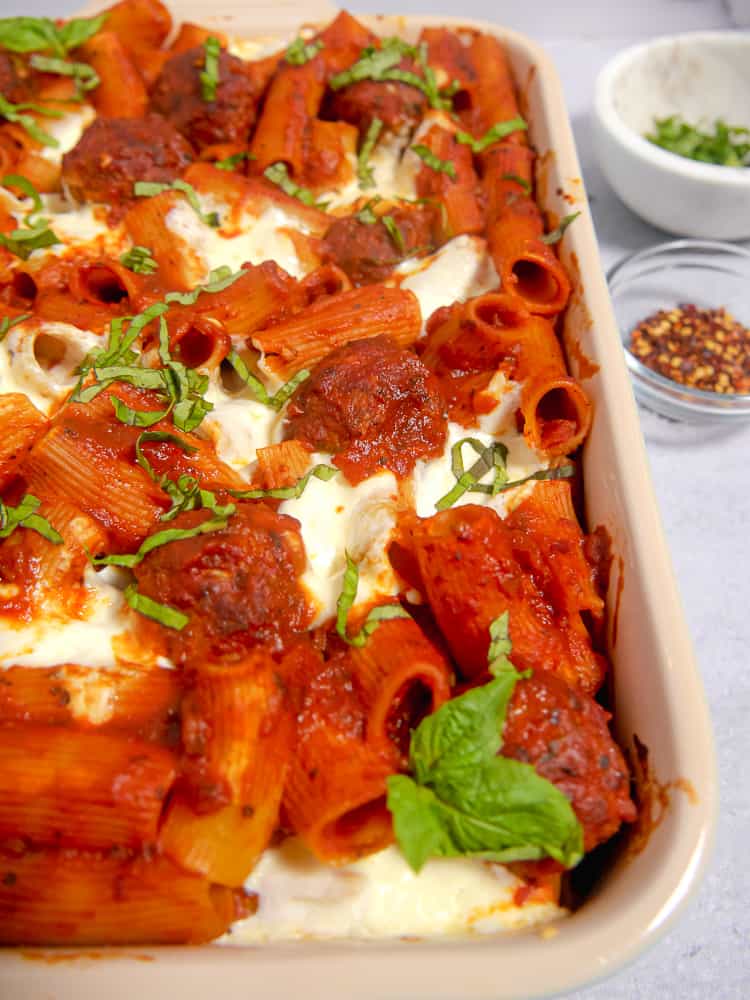 close up of rectangular baking dish with meatballs in red sauce, rigatoni, and cheese topped with fresh basil