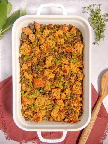 white baking dish with cornbread, sausage, apples, celery stuffing with red napkin, fresh herbs in background