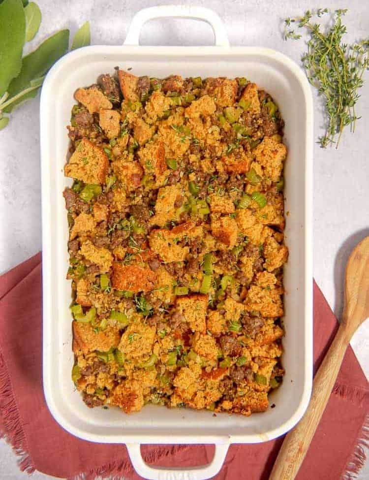 white baking dish with cornbread, sausage, apples, celery stuffing with red napkin, fresh herbs in background