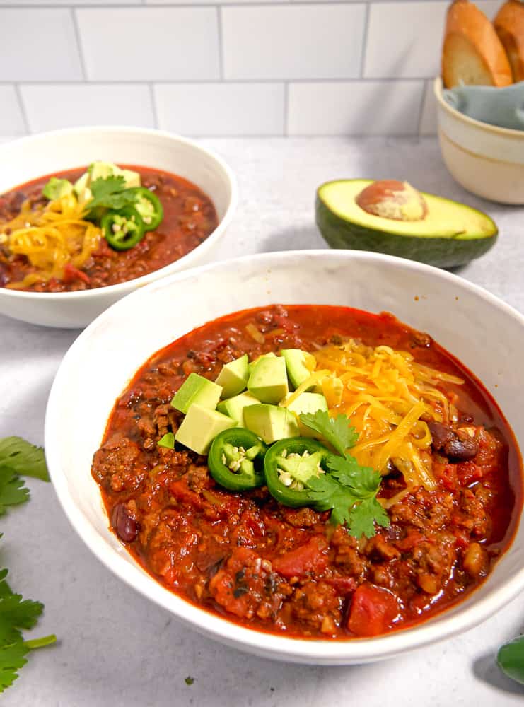 two bowls filled with beef chili, topped with cheese and avocado. Whole avocado in background with a bowl of toasted bread