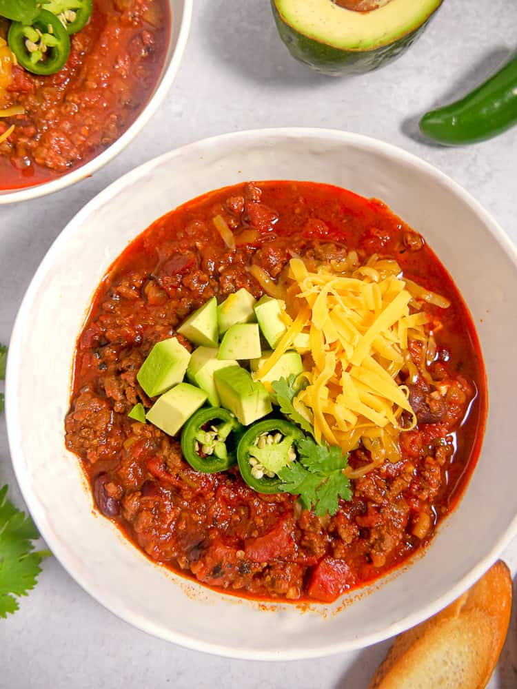 Close up of white bowl filled with beef chili topped with cheese, avocado, and jalapenos