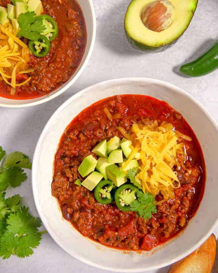 two white bowls filled with chili topped with shredded cheese, avocado, and cilantro