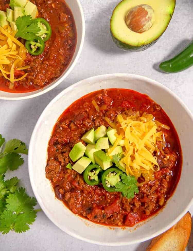 two white bowls filled with chili topped with shredded cheese, avocado, and cilantro