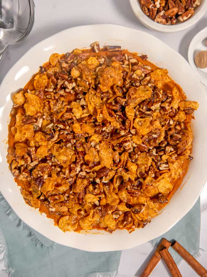 overhead photo of a sweet potato casserole with brown sugar crunchy pecan and cornflake topping in a white dish with cinnamon sticks, nutmeg, and pecans surrounding